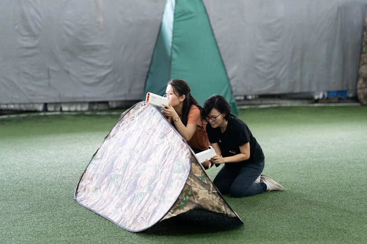 Two Laser Tag players using white laser gear behind a camouflage barrier
